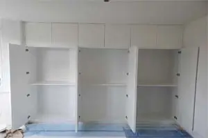 A row of three white walk-in closets with open doors. The closets have shelves, hanging rods, and drawers for organized storage.