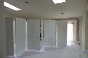 A row of five closed white doors with silver handles. Sunlight streams through a window located at the end of the hallway.